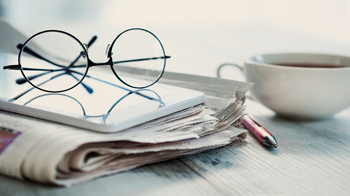 Glasses, a tablet, a newspaper, a pen, and a mug of coffee on a table