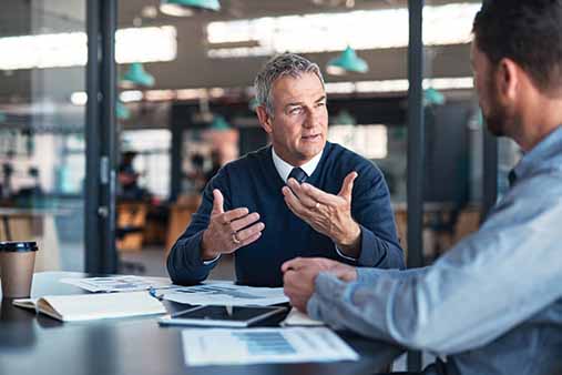 An older man explaining something to a younger man at a conference table.